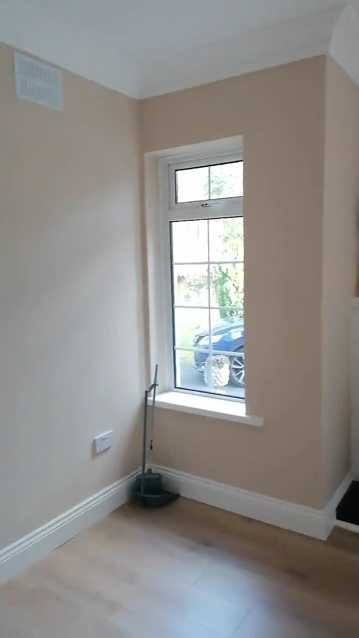  a wall in a living room with a window with white frame, magnolia walls and light brown laminate flooring and white skirting boards and white coving